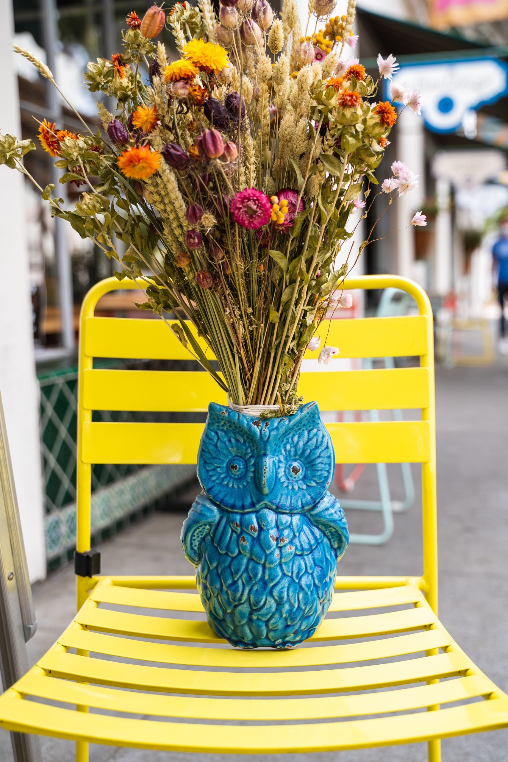 Flower arrangement in a blue owl vase sitting on a yellow chair by other stores in the background